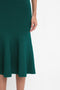 A person wearing a fitted Victoria Beckham VB Body Flared Skirt In Lurex Green with a flared silhouette hem. The image shows the lower half of their body, with one arm resting by their side.