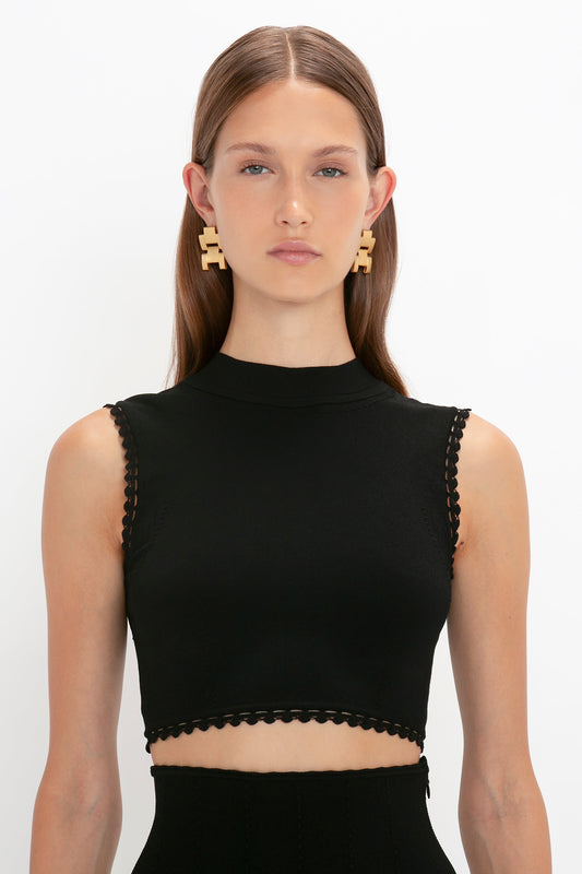 A woman with straight, light brown hair wearing a sleeveless black top and large, geometric gold earrings stands against a plain white background. The VB Body Scallop Trim Tank Top in Black by Victoria Beckham is the perfect summer essential for any wardrobe choice.