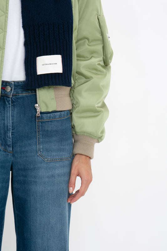 Close-up of a person wearing a green bomber jacket, blue jeans, and a blue sweater with an Exclusive Logo Patch Scarf In Navy by Victoria Beckham visible.