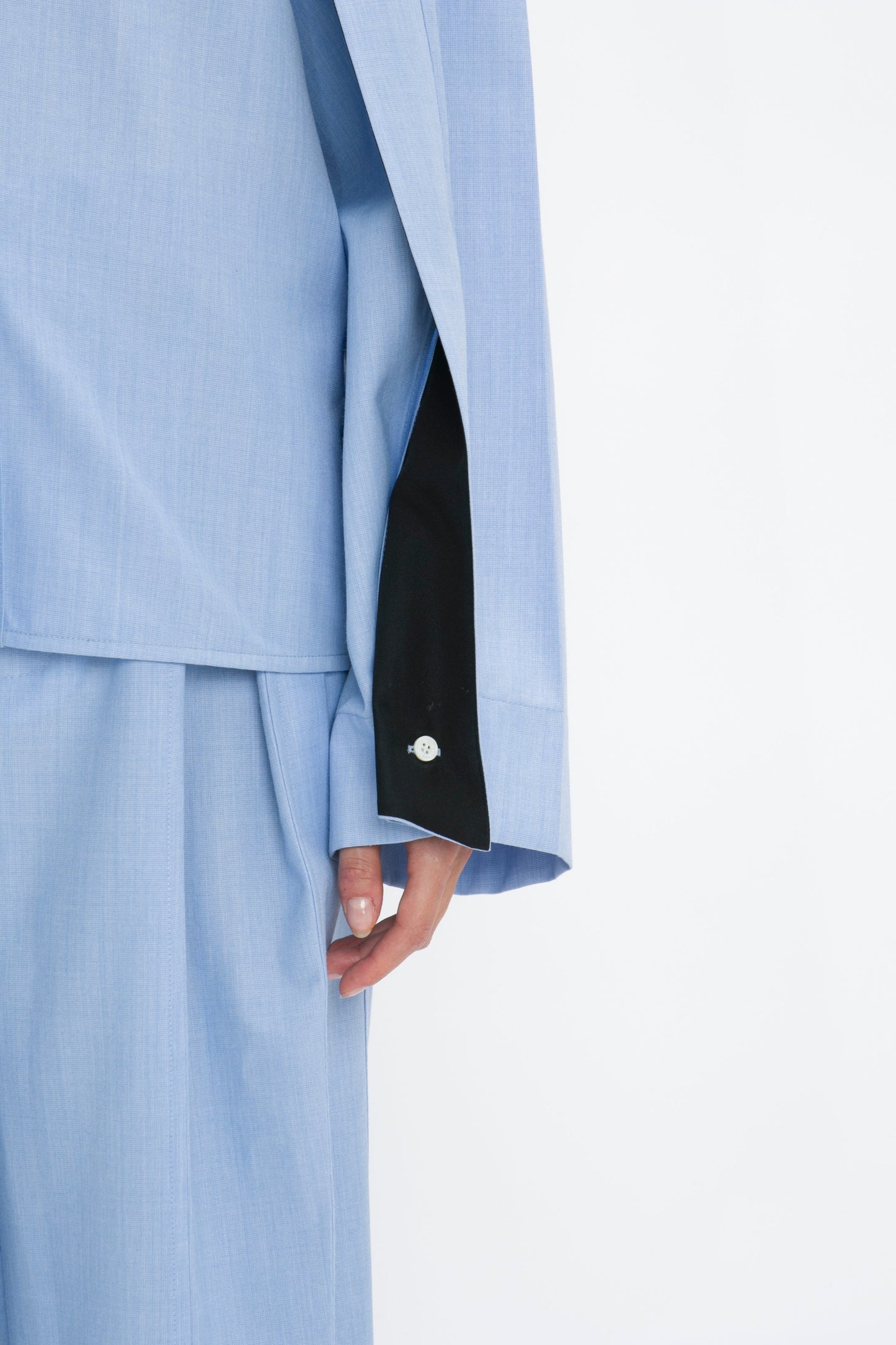 Close-up of a person wearing the Victoria Beckham Pleat Detail Raglan Shirt In Oxford Blue with raglan sleeves and a black inner lining on the sleeve. The sleeve is partially rolled up, showcasing a decorative button and part of the person's hand.