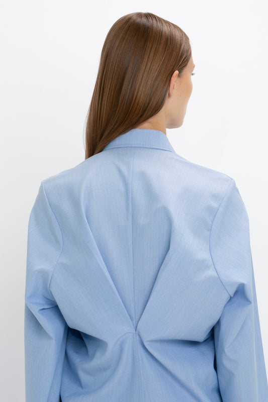 A person with long brown hair, seen from the back, wears a light blue Pleat Detail Raglan Shirt In Oxford Blue by Victoria Beckham featuring a concealed button-front closure.