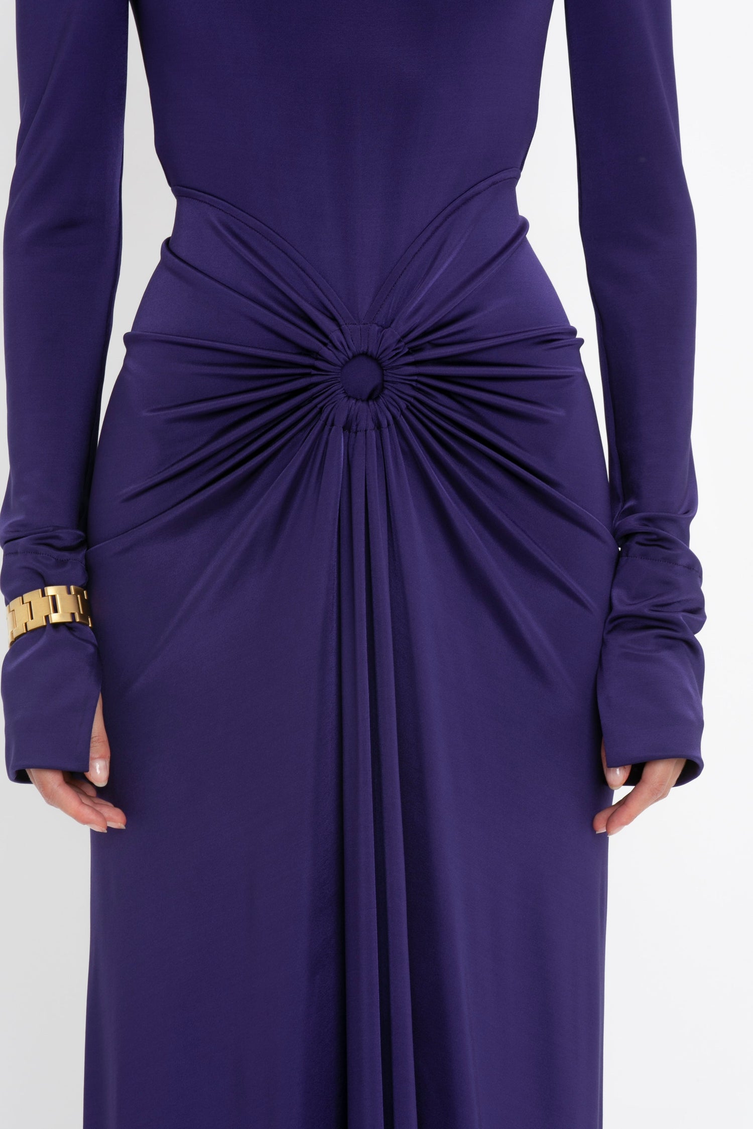 A person wearing a Victoria Beckham Long Sleeve Gathered Midi Dress In Ultraviolet made from body-sculpting stretch fabric in deep purple, featuring a central knot detail at the waist and a sensuous, playful vibe, complemented by a gold bracelet on the left wrist.