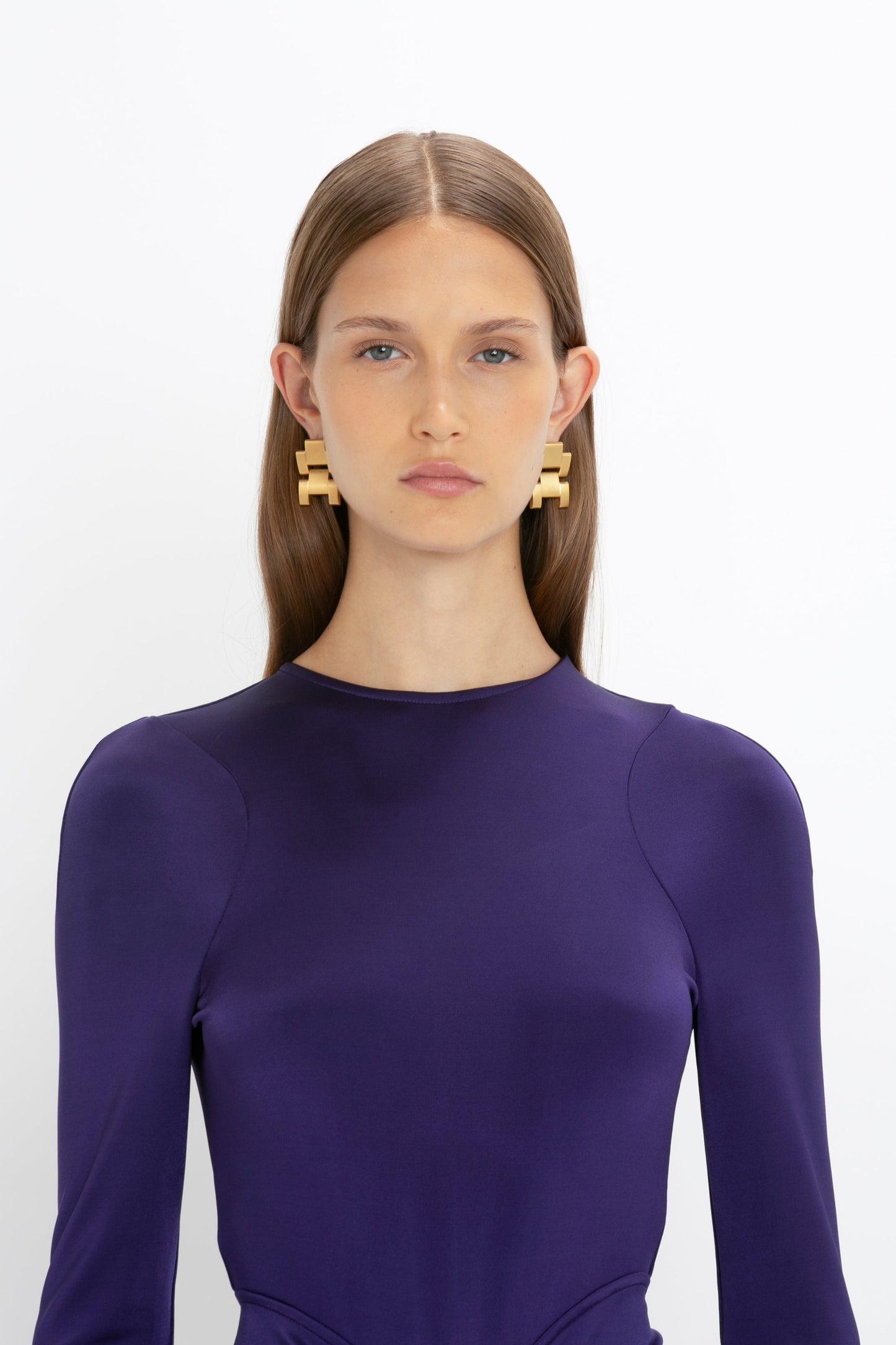 A woman with long brown hair is wearing a deep purple Long Sleeve Gathered Midi Dress In Ultraviolet by Victoria Beckham made from body-sculpting stretch fabric and large geometric earrings. She is standing against a plain white background.