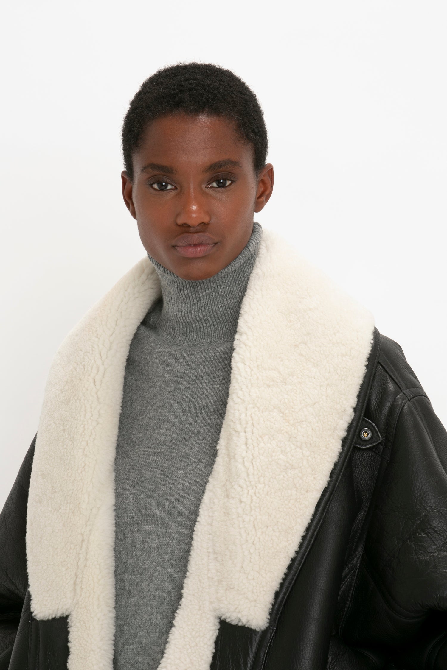 Person with short hair wearing a gray turtleneck and a black jacket with a white wool collar, standing against a plain background, giving off an effortlessly chic look. The Victoria Beckham Shearling Coat In Monochrome's oversized silhouette hints at cozy vibes perfect for the season.