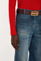 A person wearing a red long-sleeve shirt, blue denim jeans with a brown belt, and a gold buckle. The Victoria Beckham Alina High Waisted Jean In Heavy Vintage Indigo Wash are complemented by the person's right hand placed near the right pocket.