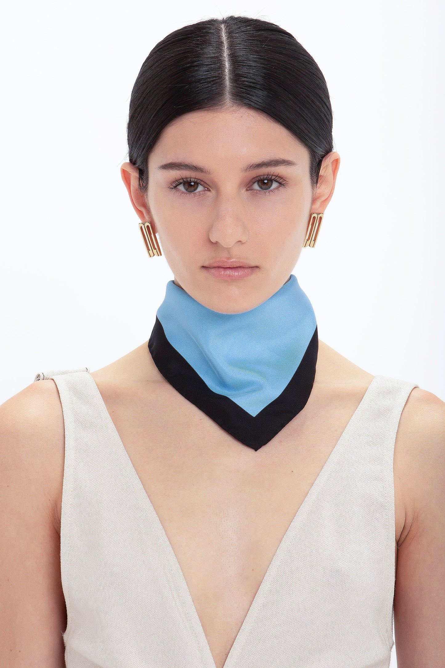 A woman with dark hair parted in the middle, wearing gold earrings and a beige top accessorized with a Victoria Beckham Colour Block Foulard In Marina scarf, against a white background.