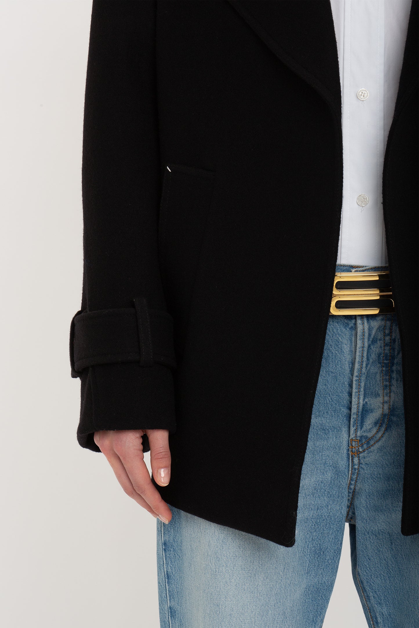 A person wearing an Victoria Beckham Oversized Pea Coat In Black and light blue jeans, showing a belt with a gold buckle, with a white shirt visible underneath.