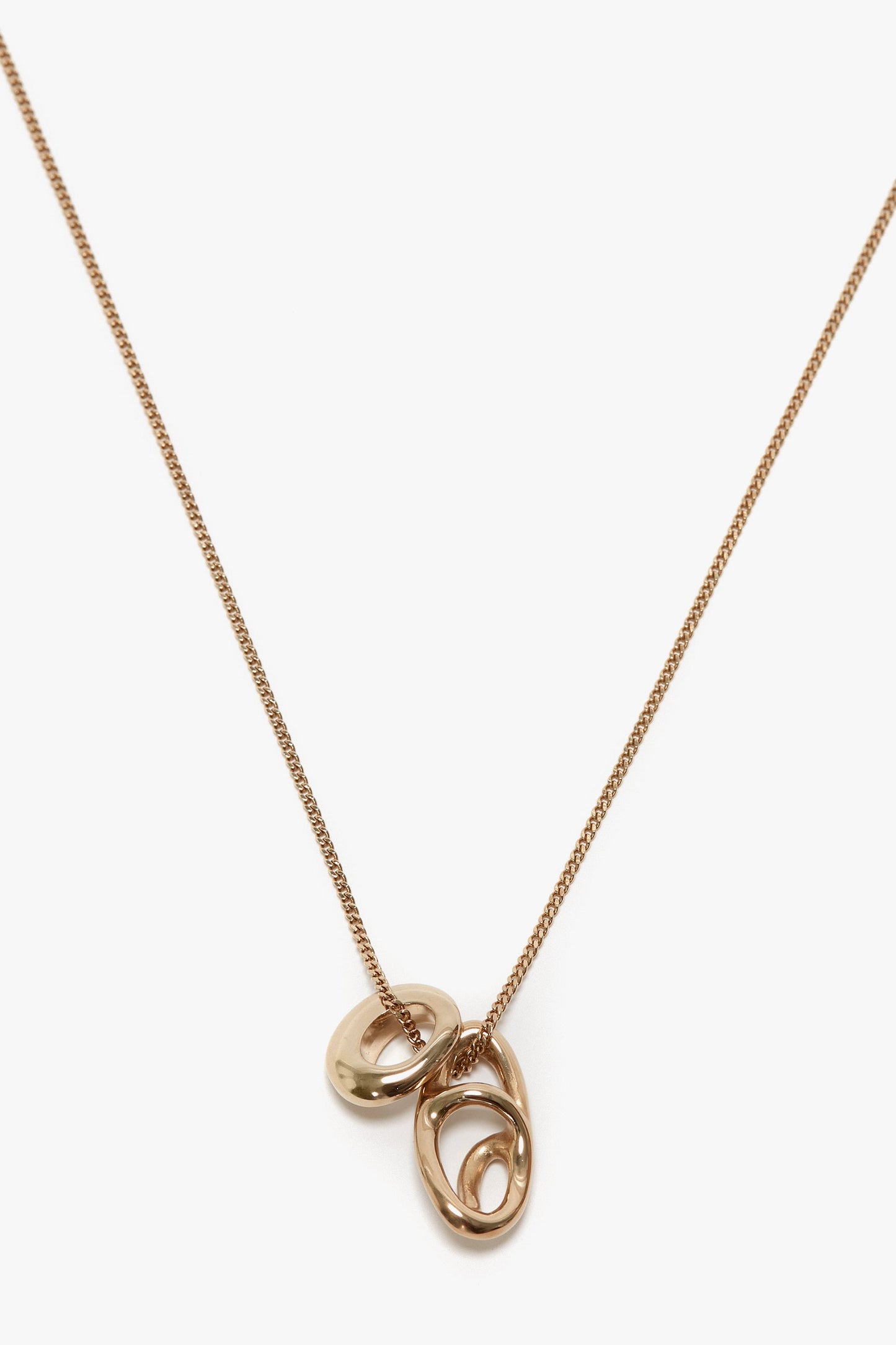 A light gold necklace with a fine chain, featuring three interlocking rings as an abstract charm pendant, displayed on a white background. Made from 100% brass. This is the Exclusive Abstract Charm Necklace In Light Gold by Victoria Beckham.