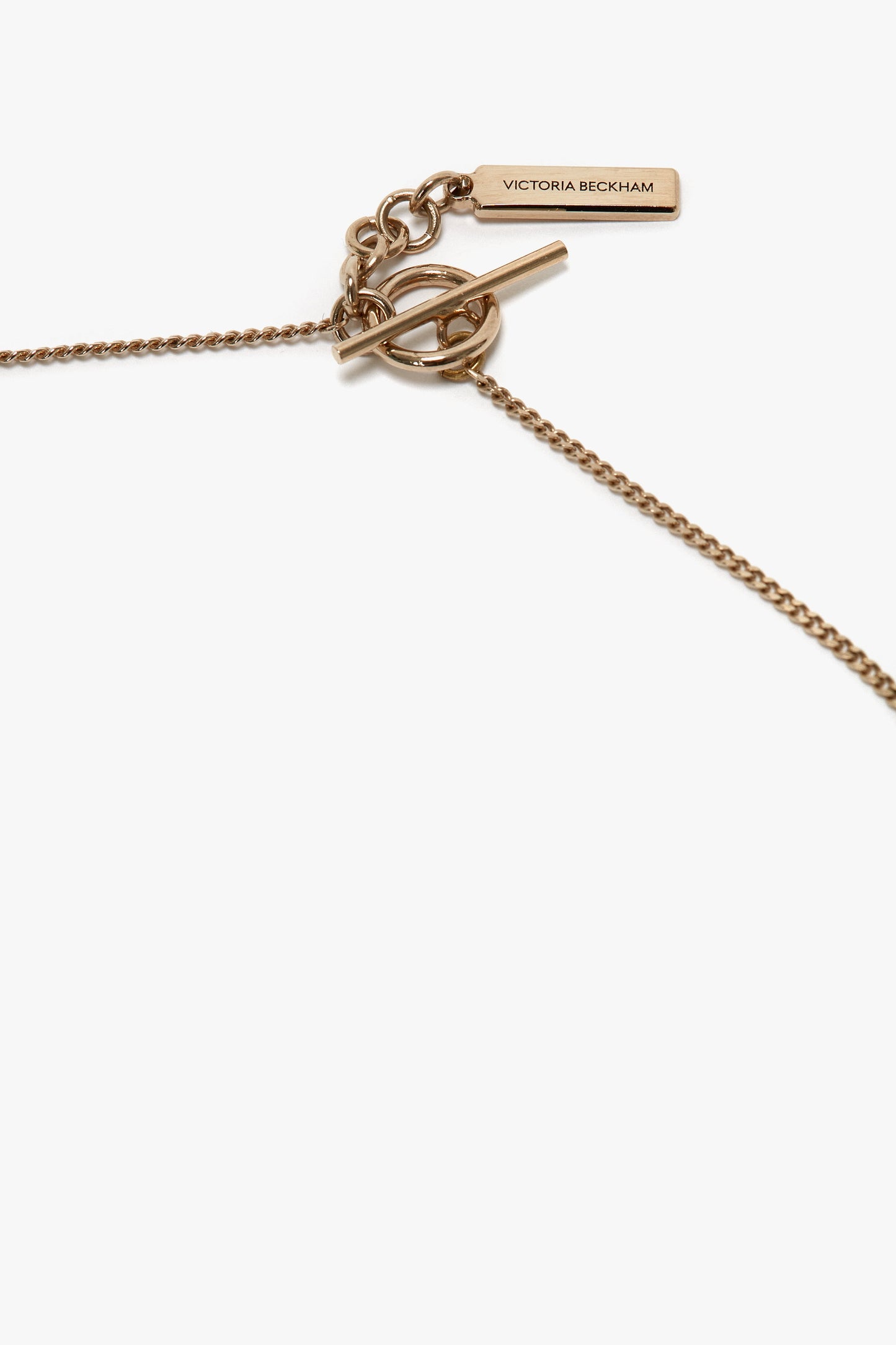 An Exclusive Body Charm Necklace In Light Gold featuring a toggle clasp and a small rectangular tag engraved with "Victoria Beckham," crafted from 100% brass.