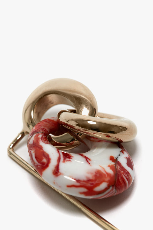 Close-up of a piece of jewelry featuring two intertwined light gold and orange metal rings and a marbled red and white *Exclusive Resin Charm Brooch In Light Gold-Orange*, made in Italy by *Victoria Beckham*.