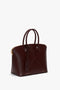 A dark brown Victoria Beckham Victoria Bag In Burgundy Leather with a structured design, double handles, an adjustable strap, a gold-toned zipper, and a small tag attached to one handle.