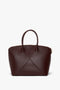 A dark brown leather Victoria Beckham Victoria Bag In Burgundy Leather with a structured shape, two short handles, and a visible seam design on the front; features an adjustable strap for versatile carrying.