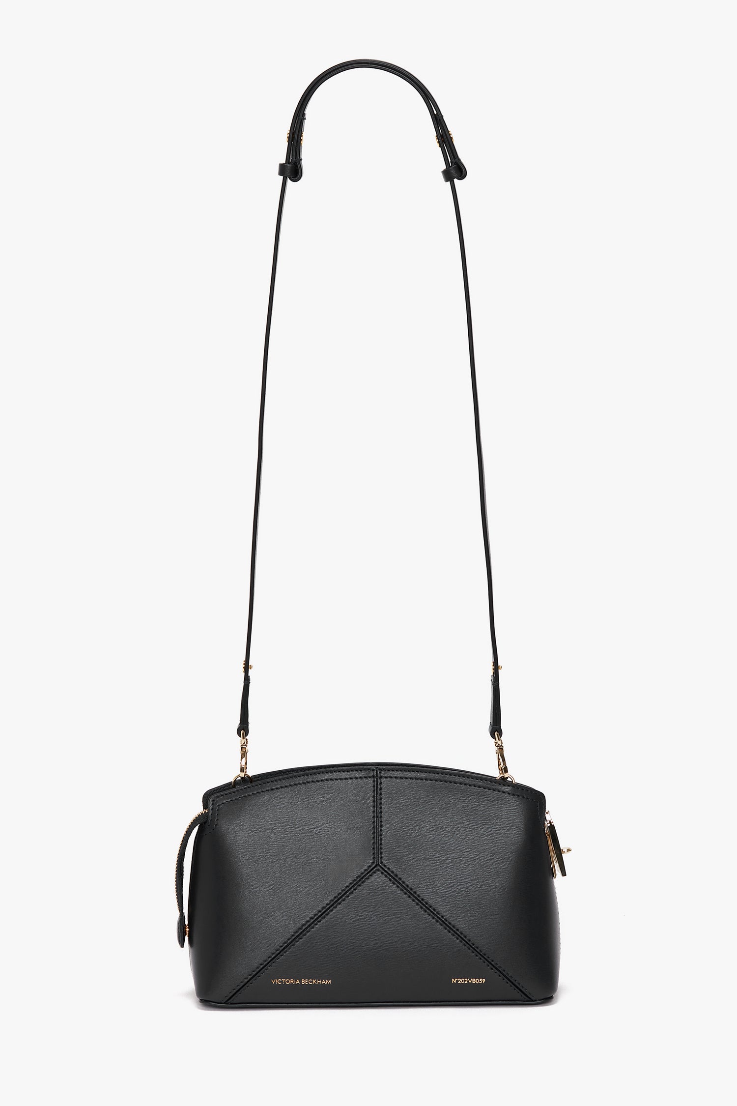 The Victoria Crossbody Bag In Black Leather is a black leather bag with an adjustable strap, gold-tone metal accents, and minimalistic stitching. Crafted from textured calf leather, it features the Victoria Beckham brand name embossed on the front.