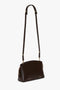 The Victoria Beckham Exclusive Victoria Crossbody Bag In Brown Leather is a small, dark brown shoulder bag made from premium calf leather, with a long adjustable strap and gold-colored hardware. It features a zippered top and a minimalist design, complete with the signature V shape.