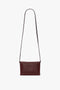 Image of a minimalist, dark red Mini Chain With Long Strap Pouch Bag In Burgundy Leather by Victoria Beckham with a long, detachable strap against a plain white background.