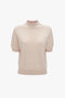 A short-sleeve, light pink sweater with a speckled pattern and a mock neck collar featuring pointelle lace stitch detailing. The collar has a contrasting red trim. This is the Short Sleeve Top In Nougat by Victoria Beckham.