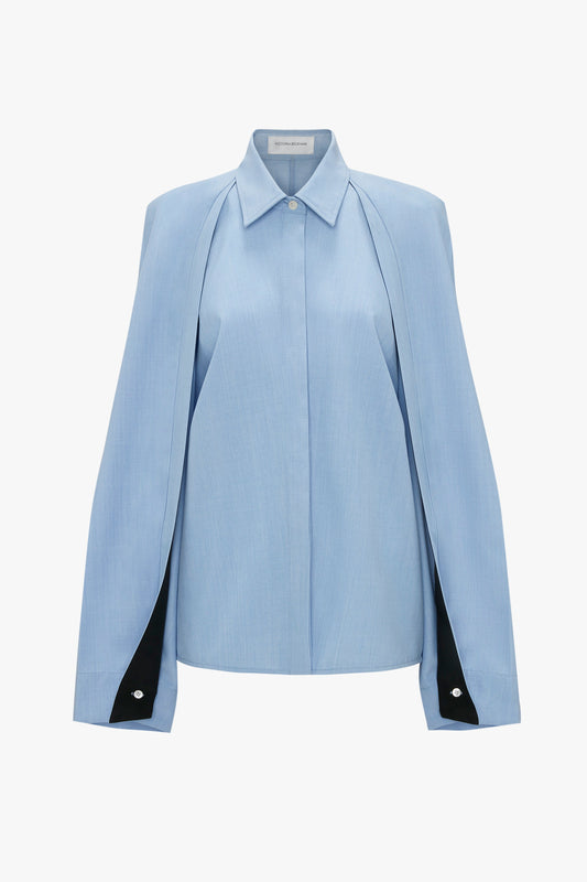 A light blue blouse with a cape-like design, long sleeves, and elegant button details at the cuffs. The Pleat Detail Raglan Shirt In Oxford Blue by Victoria Beckham also features raglan sleeves, adding a modern twist to its timeless style.