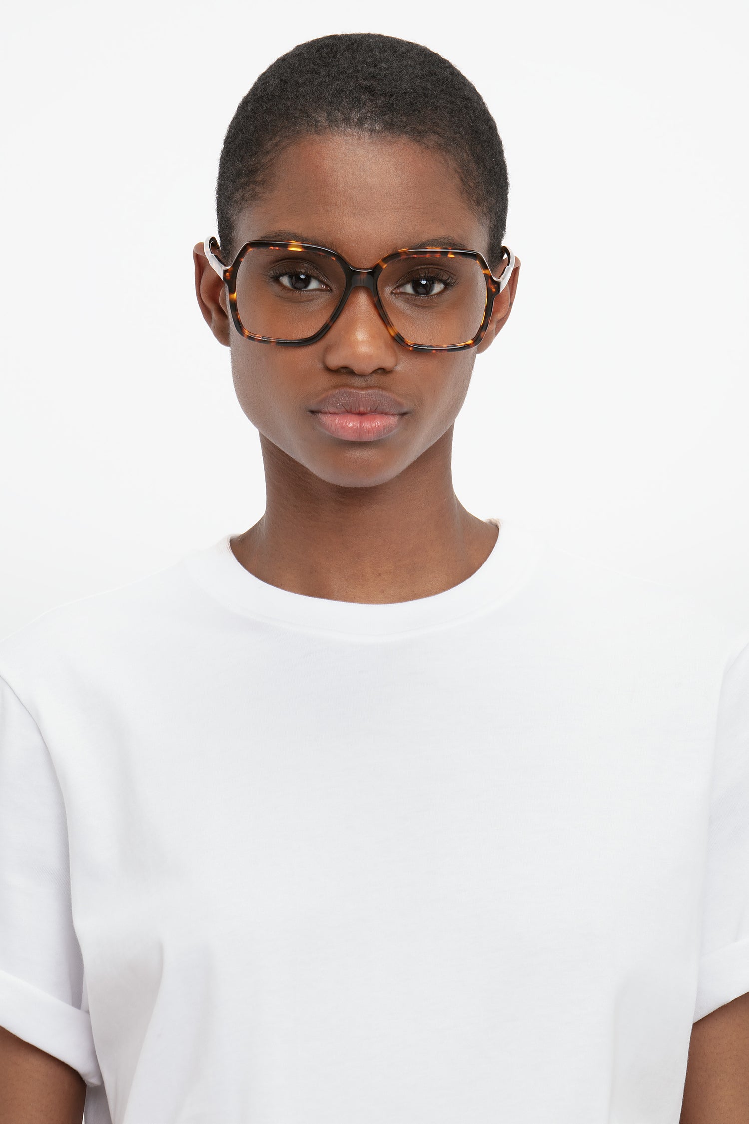 A person with short hair wearing large, Guilloche Frame Opticals In Dark Havana by Victoria Beckham and a white t-shirt looks directly at the camera against a plain white background.