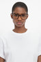 A person with short hair wearing large, Guilloche Frame Opticals In Dark Havana by Victoria Beckham and a white t-shirt looks directly at the camera against a plain white background.