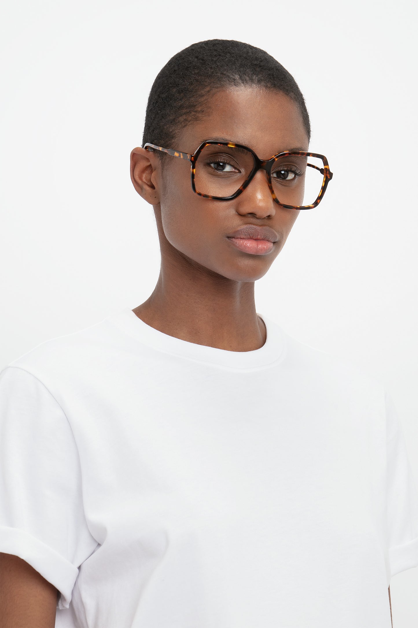 Person wearing large, Guilloche Frame Opticals In Dark Havana by Victoria Beckham and a plain white t-shirt, looking straight at the camera against a plain background.