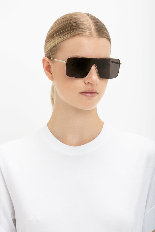 A person with blonde hair pulled back, wearing a white shirt and large, Rectangular Visor In Gold-Smoke from Victoria Beckham, exudes contemporary elegance against a white background.