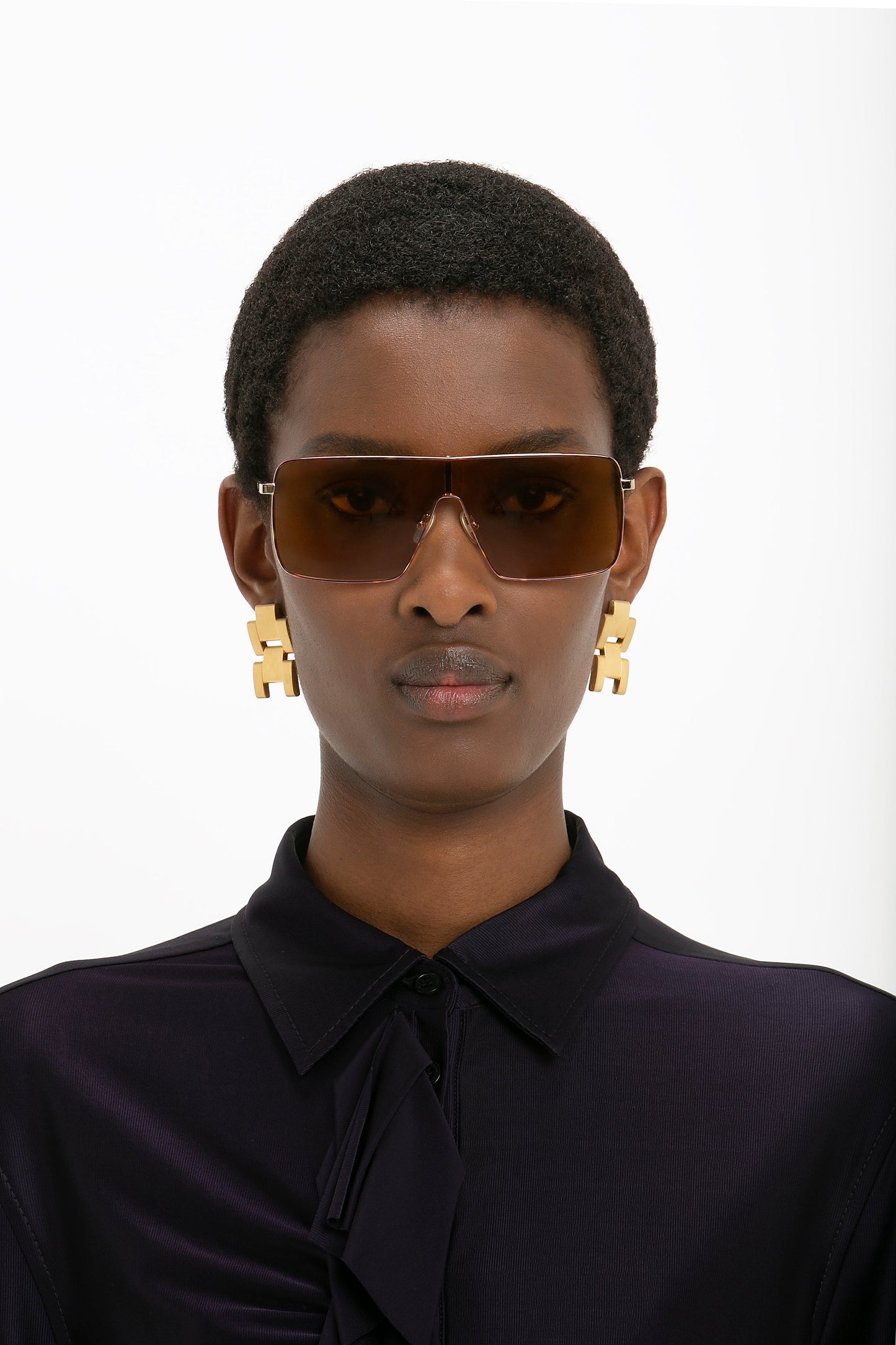 A person wearing Rectangular Visor Gold-Framed Sunglasses by Victoria Beckham, gold geometric earrings, and a dark-colored top against a white background.