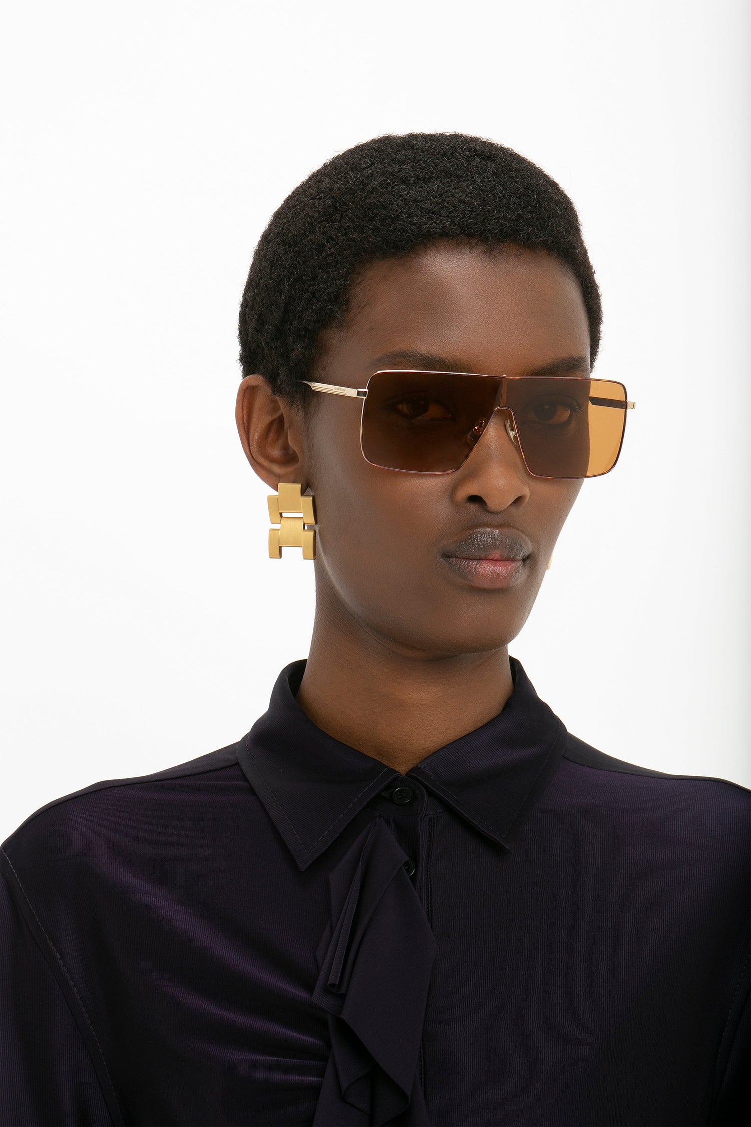 Person wearing Victoria Beckham Rectangular Visor Gold-Framed Sunglasses, gold geometric earrings, and a dark shirt with a high-neck collar.