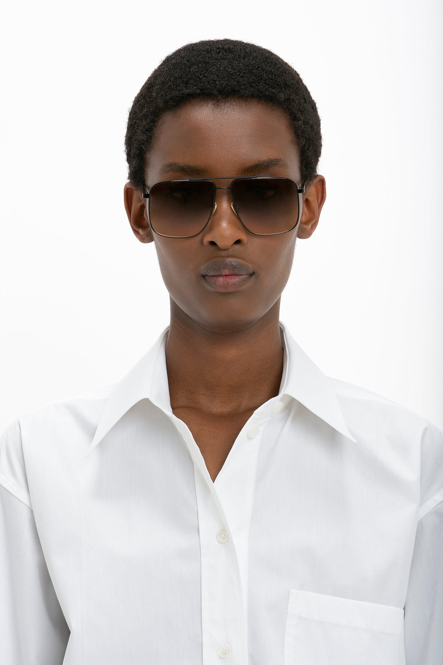 A person wearing Victoria Beckham Double Bridge V-Line Navigator In Gold-Black-Brown sunglasses and a white button-up shirt looks directly at the camera against a plain white background.