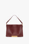 A Jumbo Chain Pouch Bag In Bordeaux by Victoria Beckham with a single shoulder strap, gold hardware, and a rectangular decorative clasp at the bottom. The bag also features a detachable strap for added versatility.