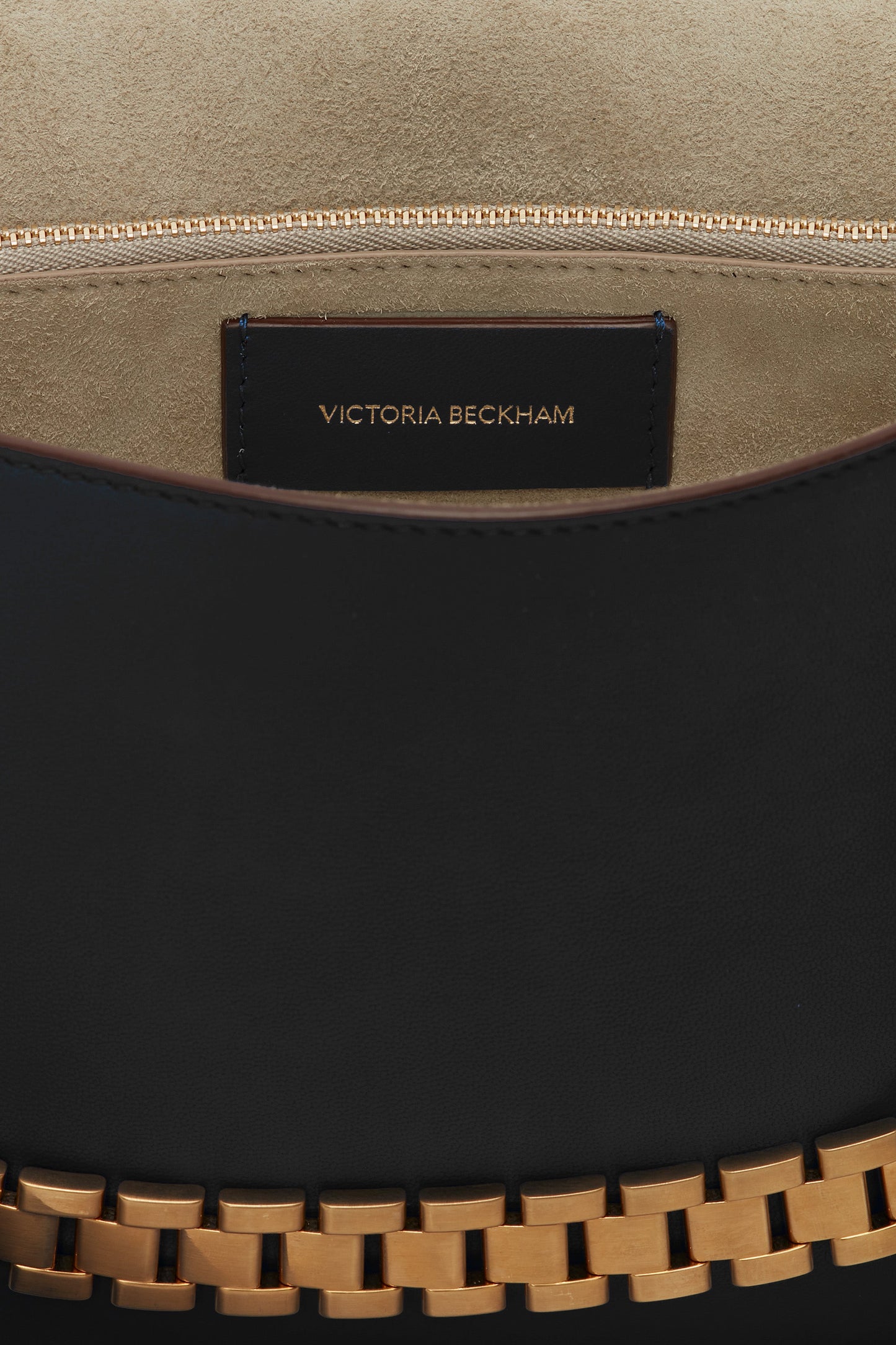 Close-up view of the interior of a black Victoria Beckham Chain Pouch Bag with Strap In Black Leather, crafted from Nappa leather, showing the brand's name on a leather tag near a zippered pocket and a detachable handle with a golden chain detail at the bottom.